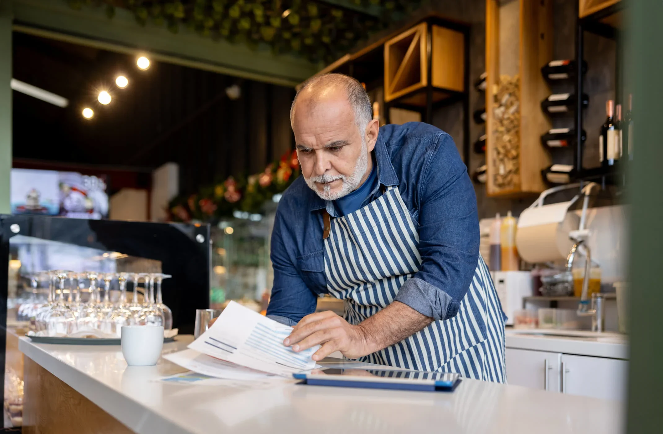 A business owner in a striped apron stands in his kitchen reading his utility bills
